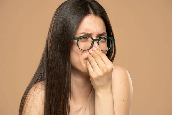 Portrait of frustrated woman pinching nose with disgust on his face due to bad smell isolated over beige background.