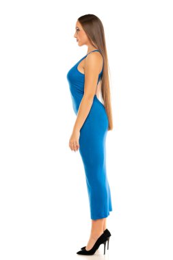 Beyaz arka planda duran, uzun şeffaf mavi elbiseli genç bir kadının yan görüntüsü.