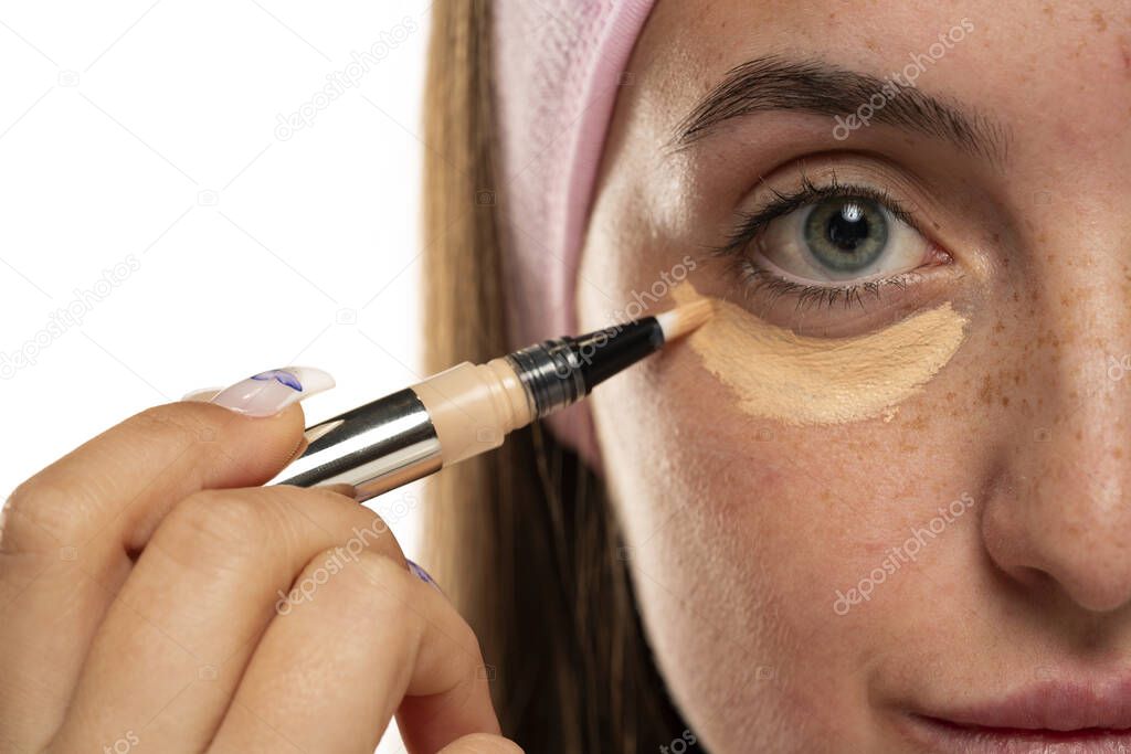Young woman applyes concealer under her eyes on white background