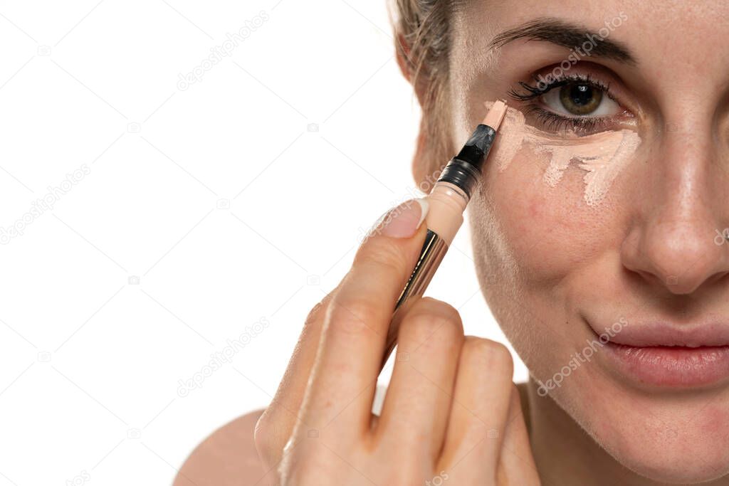 Half portrait of a young woman applies concealer under her eyes on white background.