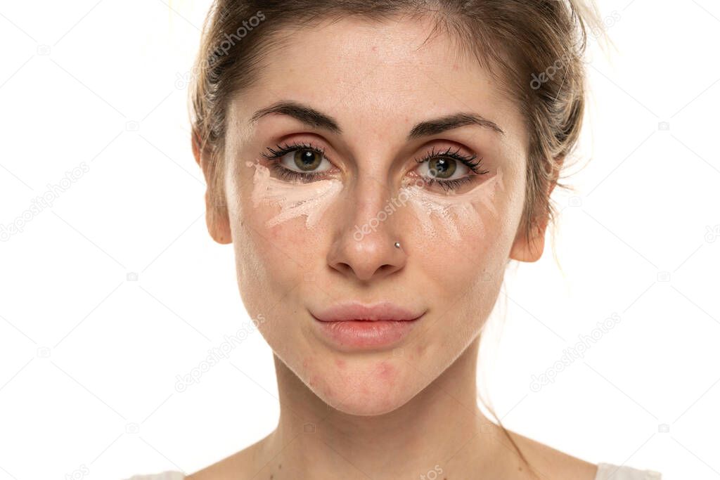 Young woman posing with concealer under her eyes on white background.