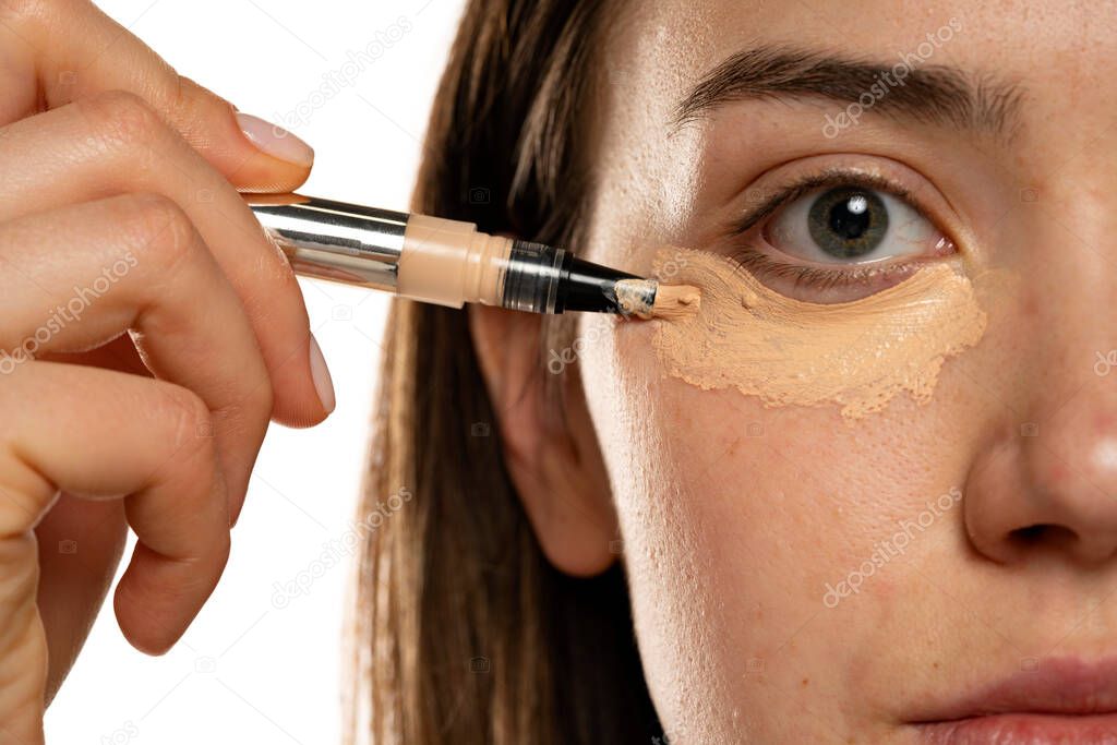 Close up of a young women applying concealer under her eyes on white background