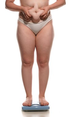 Fat belly woman standing on a scale and squeezing a fat roll on her belly on white background.