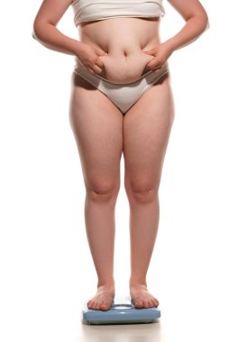 Fat belly woman standing on a scale and squeezing a fat roll on her belly on white background.