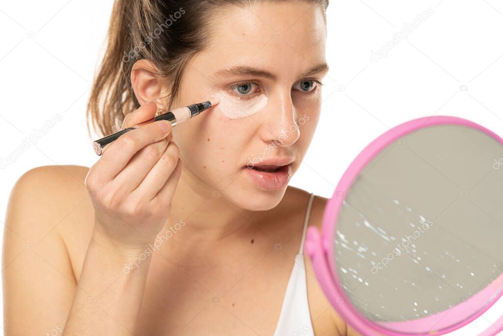 Young woman applyes concealer under her eyes on white studio background.