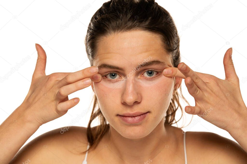 Young woman applyes concealer under her eyes on white background.
