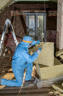 Tavan arası yenileme ve yalıtım konsepti. Mineral yün panelleri kesen üniformalı bir işçi.