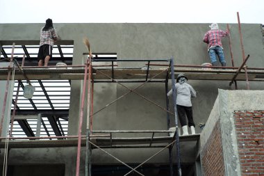 İnşaat alanındaki beton duvarları sıva yapmak için yüksek yapı iskelesinde duran işçilerin arka görünümü