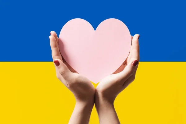 Women hands holding heart shape paper on Ukraine flag color background. Stay with ukraine symbol. Hand heart love gesture with ukrainian flag background.