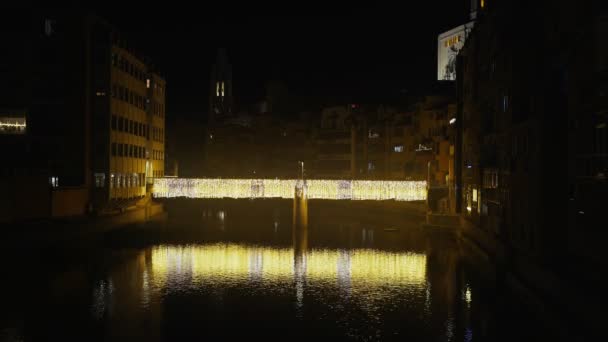 Beautiful illuminated bridge over the river in the old town. Festive city illumination. Yellow lights are reflected in ripples of water. Night view of the medieval city. Girona. Catalunya. Spain. — стокове відео