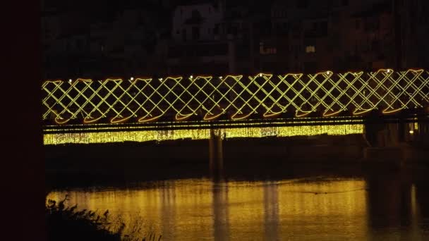 Beautiful illuminated bridge over the river in the old town. Festive city illumination. Yellow lights are reflected in ripples of water. Night view of the medieval city. Girona. Catalunya. Spain. — стокове відео