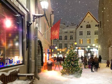 Ortaçağ şehir caddesinde akşam vakti aydınlatılmış Noel ağacı, vitrinlerde fener ışığı bulanık, gece gökyüzü dolunayı insanlar Tallinn 'deki eski kasaba meydanında karlı havada yürüyor. 