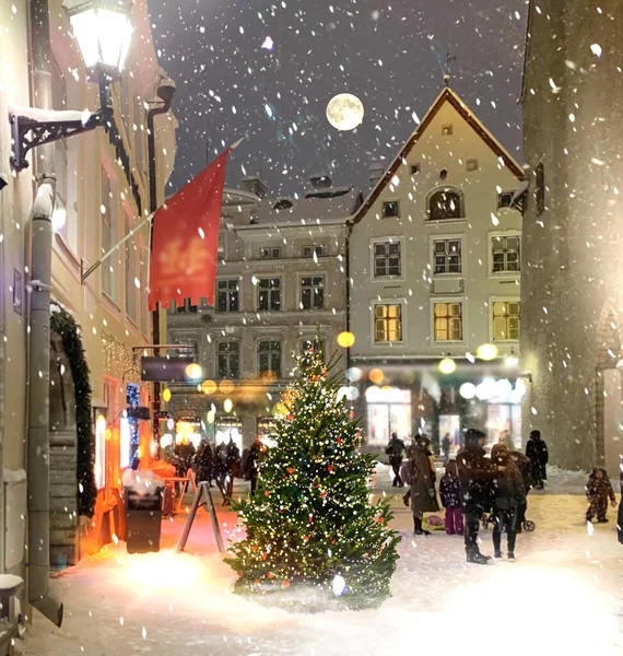 中世の街の通りの夜の冬に照らされたクリスマスツリー 店の窓のつるでぼやけた提灯の光 夜の空満月の人々タリン旧市街ホール広場市場で雪の天気の中を歩く人々 — ストック写真