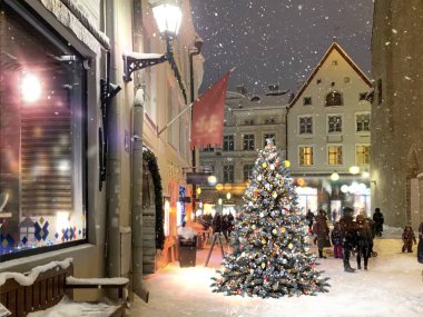 Ortaçağ şehir caddesinde akşam vakti aydınlatılmış Noel ağacı, vitrinlerde fener ışığı bulanık, gece gökyüzü dolunayı insanlar Tallinn 'deki eski kasaba meydanında karlı havada yürüyor. 