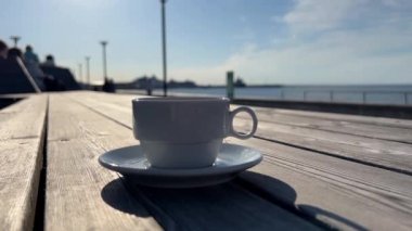 Ufukta deniz manzarası ve mavi gökyüzü olan plaj kafesindeki ahşap masada bir fincan kahve.