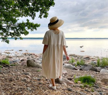  Sahilde, saman şapkalı, vahşi plajda beyaz elbiseli, kıdemli zarif kadın deniz ve gökyüzünü seyrediyor. Romantik yaz arkaplanı.