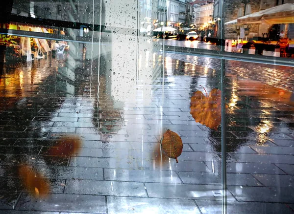 rain in  city street, people silhouette with umbrellas ,modern and vintage building ,Rainy weather season ,Autumn leaves on window,wet drops ,night  blurred light  reflection cold urban  defocus   background