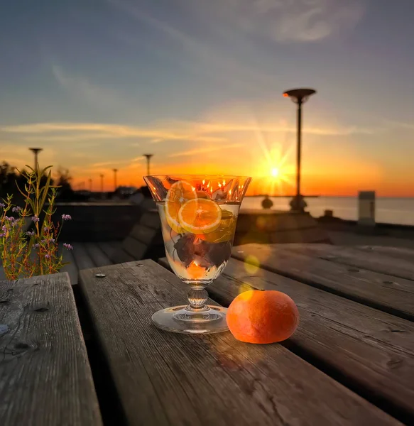 glass of orange water and mandarine  freshness  on wooden table  and flowers on promenade at sunset in sea people relax summer