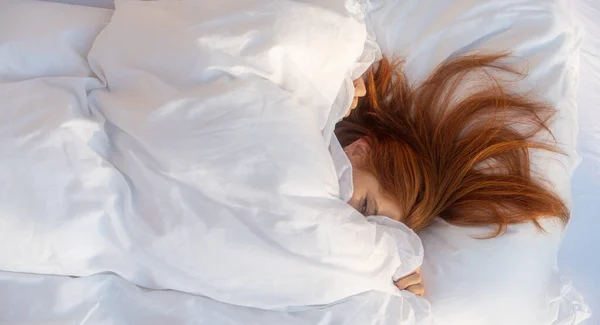 Çekici, genç, seksi, kızıl saçlı, yüzü neredeyse tamamen yastıklarla kaplı, bir gözü dışarıda, yatak odası güneşinde taze, yumuşak, beyaz bir yatakta yatıyor,