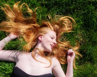 Genç, seksi, kızıl saçlı bir kadının güzel portresi, ilkbaharda uzanıyor, yaz güneşi, gevşiyor, gülümsüyor, gülüyor, yeşil çayır çimlerin üzerinde, başındaki uzun kızıl saçlar, kopyalama alanı.