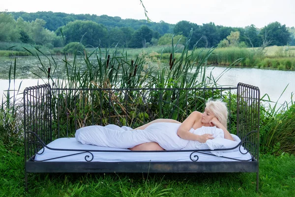 attractive, young, sexy, blonde woman lying naked covered with white duvet in bed sleeping in the nature in front of a lake with reeds and bulrushes.