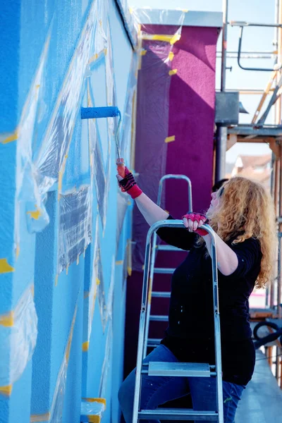 adult mature redhead painter woman with curly red hair and sunglasses, stands on ladder and paints artistic polygonal color patch in blue color paint on the house wall, with blue painter roller.