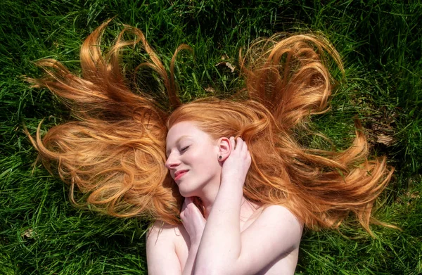 Top view beautiful portrait of young sexy redhead woman, lying happy in the spring, summer sun, relaxing, smiling, laughing, on green meadow grass, the long red hair around the head, copy space