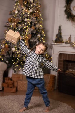 Evde Noel hediyesi olan mutlu küçük bir kız. Küçük kız yılbaşı ağacının yanında ışık arka planında hediyesini tutuyor. Çocuk oturma odasında Noel hediyesini açıyor. Yeni yılı ve aile tatillerini kabul et. 