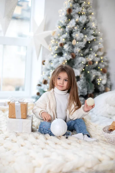 Happy little smiling girl with gift on Christmas Eve sit on bed. Child opens New year gift near christmas tree with glowing lights garland. Kid play in bed with xmas balls and toys in bedroom at home