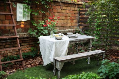 Evdeki arka bahçede yemek masası. Avluda ya da balkonda rahat bir yer. Teras, tuğla duvar ve bitkilerle dolu, masa ve ahşap bank. Bahçe yazın veranda, ahşap bahçe mobilyası ve barbekü.