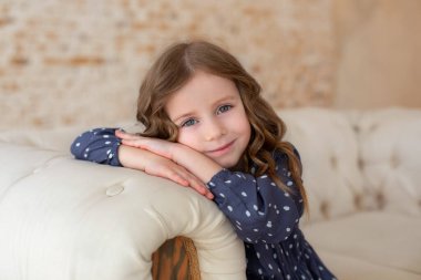 Güzel genç kız kanepede dinleniyor ve kameraya bakıyor. Oturma odasında kanepede oturan gülümseyen küçük kız. Büyük gözleri ve kıvırcık saçları olan ilham verici küçük bir kız. Çocukluk konsepti. Rüya gören çocuk