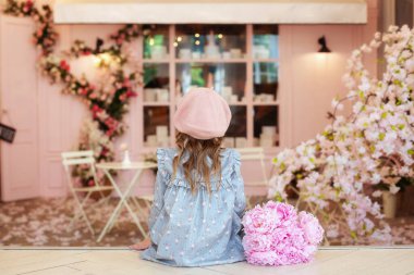  Fırın vitrininin önünde oturan genç kız. Kıvırcık saçlı, elbiseli, bereli, şakayık buketli, klasik kafede oturan, arkadan manzaralı mutlu bir kız. Çocukluk konsepti. Fransız tarzı