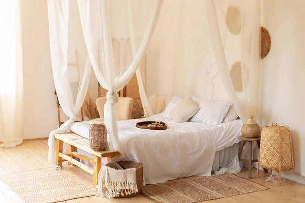 hygge decor at home. interior white bedroom in boho  style with coffe table,straw lamp and comfortable bed with pillows, copy space. bed with flowing white curtains. Cozy scandinavian decoration room