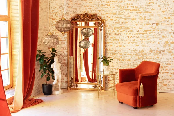 Modern living room in French style. Stylish interior with Burgundy comfortable armchair, green house plants in pots, coffee table, vintage gold mirror and crystal chandeliers against a brick wall.