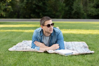 Young man in sunglass relaxing and reading book while lying on lawn. Happy Man Lying On Green Grass and study remotely. Adult man rest in the park. Smiling man working on outdoor