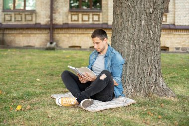 Young man relaxing and reading book while sit on lawn. Happy Man sitting On Green Grass and study remotely. Adult student reading in the park. Enjoying student life. smiling man working on outdoor 