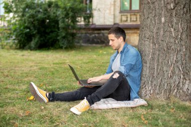 Young man with laptop rest in grass at university campus. Business man sit on lawn work outdoors on computer in a park. Student man on lesson with laptop. education and remote working concept