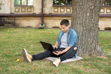 Young man with laptop rest in grass at university campus. Business man sit on lawn work outdoors on computer in a park. Student man on lesson with laptop. education and remote working concept
