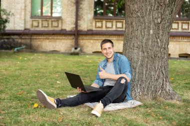 Portrait of positive man hold laptop show thumb up on grass at university campus. Business man sit on lawn work outdoors on computer in a park. Education and remote working concept. 