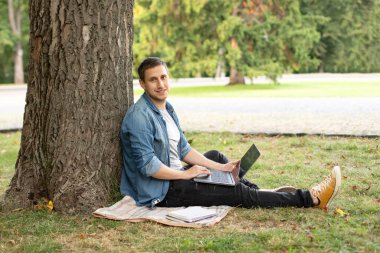 Portrait of positive man hold laptop show thumb up on grass at university campus. Business man sit on lawn work outdoors on computer in a park. Education and remote working concept. 