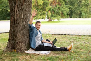 Portrait of positive man hold laptop show thumb up on grass at university campus. Business man sit on lawn work outdoors on computer in a park. Education and remote working concept. 