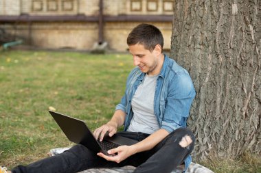 Young man with laptop rest in grass at university campus. Business man sit on lawn work outdoors on computer in a park. Student man on lesson with laptop. education and remote working concept