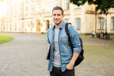 Successful student is standing with backpack and laptop behind park near campus. Smile young man standing against university on the background. Education, university, college, studying, course concept