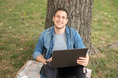 Young man with laptop rest in grass at university campus. Business man sit on lawn work outdoors on computer in a park. Student man on lesson with laptop. education and remote working concept