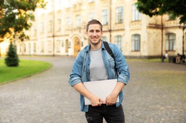 Successful student is standing with backpack and laptop behind park near campus. Smile young man standing against university on the background. Education, university, college, studying, course concept
