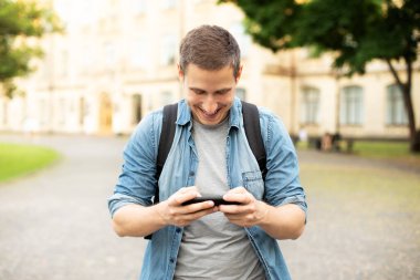 Happy man using cell phone gadget for checking email message, smiling male blogger browsing website in social media. Smiling Guy with backpack play in phone on video game in park
