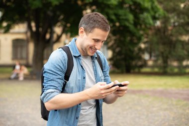Smiling Guy with backpack play in phone on video game in park. Happy man using cell phone gadget for checking email message, smiling male blogger browsing website in social media. 