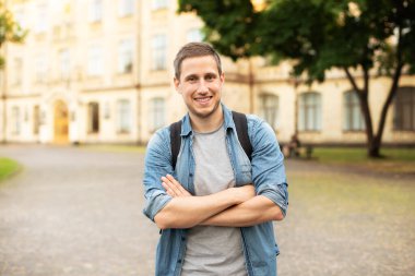 Successful student is standing with backpack near campus on the university background. Smiling young man with bag standing in park. Education, university, college, studying, course concept