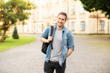 Successful student is standing with backpack near campus on the university background. Smiling young man with bag standing in park. Education, university, college, studying, course concept