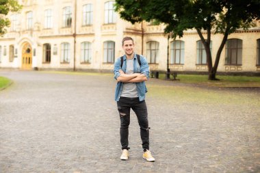 Successful student is standing with backpack behind park near campus. Smile young man standing against university on the background. Education, university, college, studying, course concept
